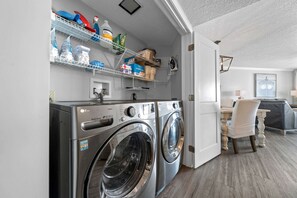 A laundry area with two front-loading LG washers, shelves above holding cleaning supplies, and a dining area with a table and chairs. - Beach Front 7th Floor Views Pools Hot Tub (Orange Beach)