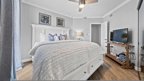 A bedroom with a bed, a ceiling fan, a TV on a wooden stand, and artwork on the wall. - Beach Front 7th Floor Views Pools Hot Tub (Orange Beach)