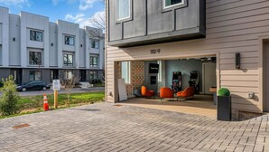 Un bâtiment moderne doté d’un toit plat, d’une entrée bien visible et d’un petit espace extérieur aménagé avec des chaises orange.