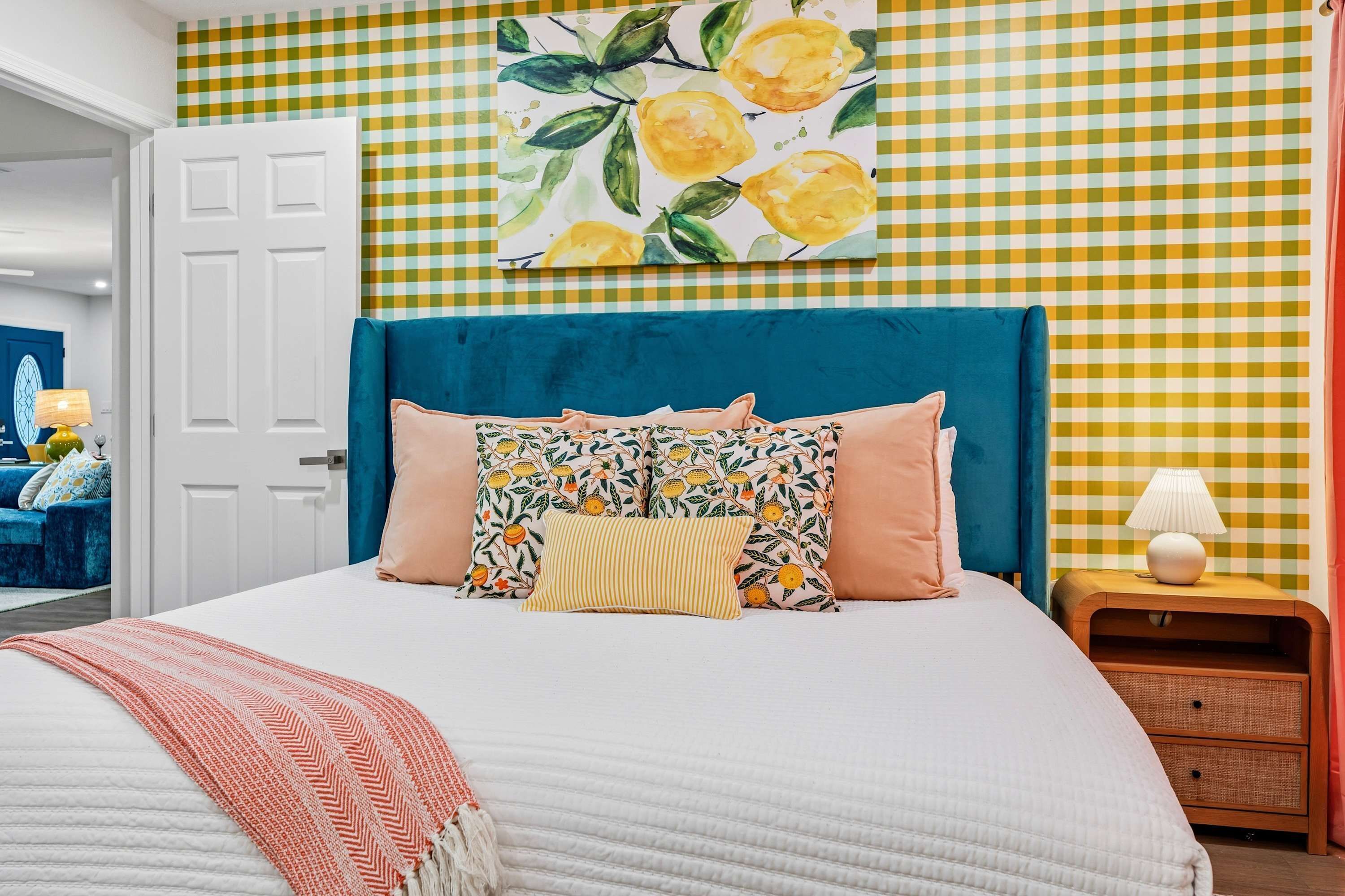 Un dormitorio con pared de cuadros, una cama con muchas almohadas, una mesita de noche con lámpara y una puerta a otra habitación.