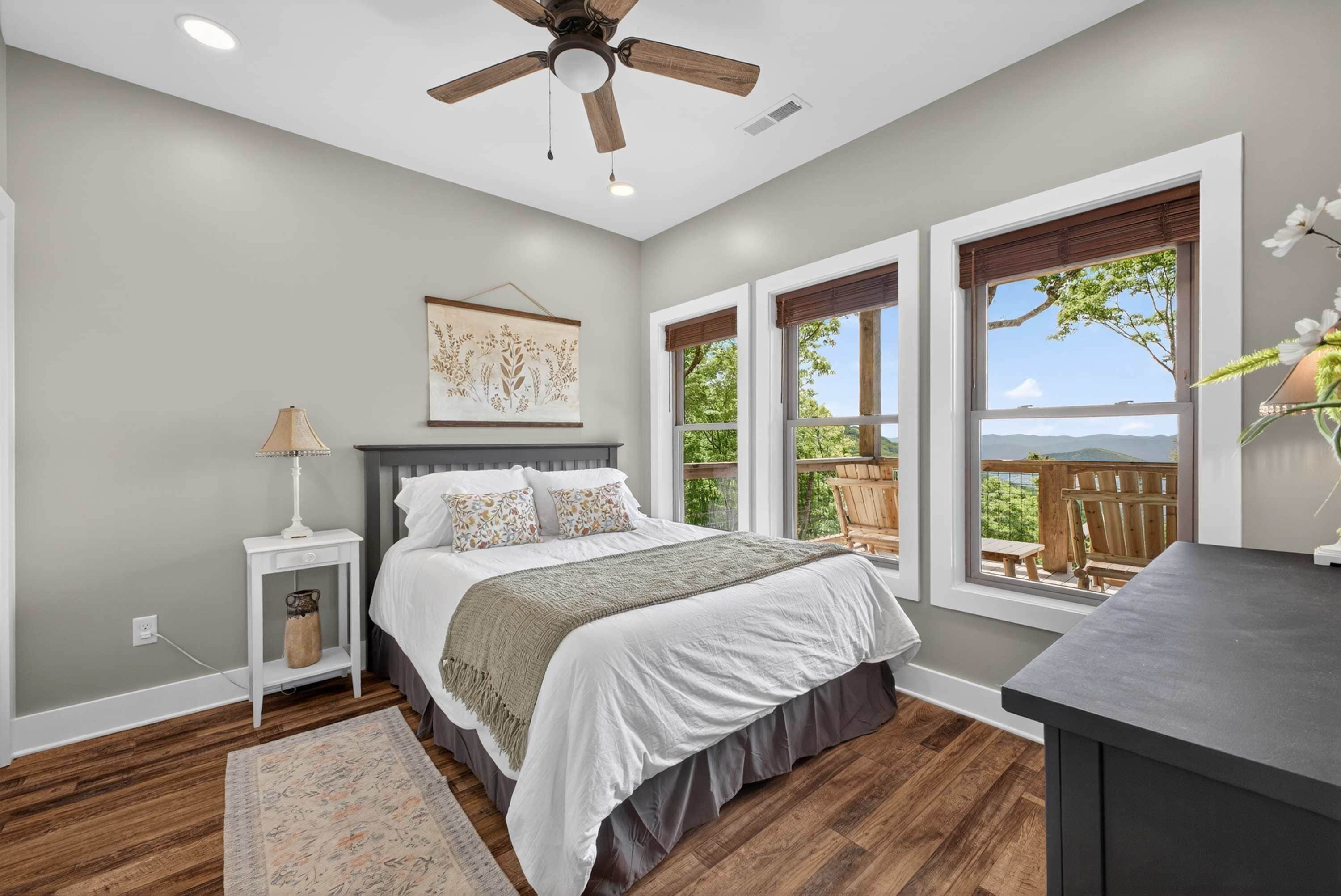 A bedroom with a bed, a nightstand, a lamp, a ceiling fan, and large windows with a view of the outdoors.