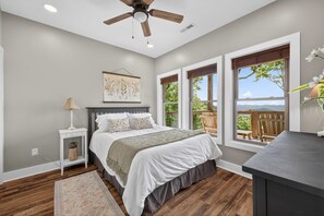 A bedroom with a bed, a nightstand, a lamp, a ceiling fan, and large windows with a view of the outdoors.
