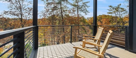 Una terraza de madera con barandilla de metal, una mecedora de mimbre y vistas a árboles otoñales.