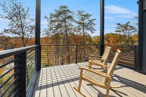 A wooden deck with a metal railing, a wicker rocking chair, and a view of autumn trees. - Luxury 3 Br Cabin Indoor Pool Hot Tub Fire Pit (Sevierville)