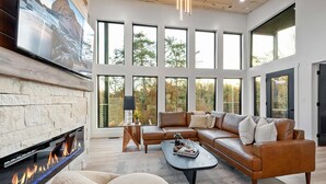 A modern living room with a fireplace, large windows, a leather sofa, and a coffee table. - Luxury 3 Br Cabin Indoor Pool Hot Tub Fire Pit (Sevierville)