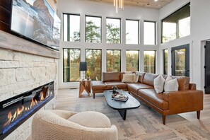 A modern living room with a fireplace, large windows, a leather sofa, and a coffee table. - Luxury 3 Br Cabin Indoor Pool Hot Tub Fire Pit (Sevierville)
