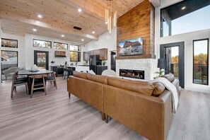 A spacious living area with a fireplace, a dining table with chairs, a sofa, and a kitchen in the background. - Luxury 3 Br Cabin Indoor Pool Hot Tub Fire Pit (Sevierville)