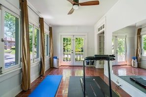 Une pièce spacieuse dotée d’un ventilateur de plafond, de grandes fenêtres avec des rideaux, d’un tapis de course et de tapis de sol pour les exercices.