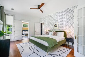 Ein Schlafzimmer mit einem Bett, einem Deckenventilator, strukturiertem Wandpapier und Blick durch eine geöffnete Tür.