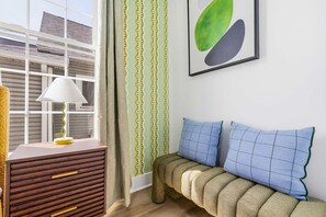 Una habitación con un aparador de madera, una lámpara, un sofá con cojines azules, una cortina estampada y un cuadro abstracto enmarcados.