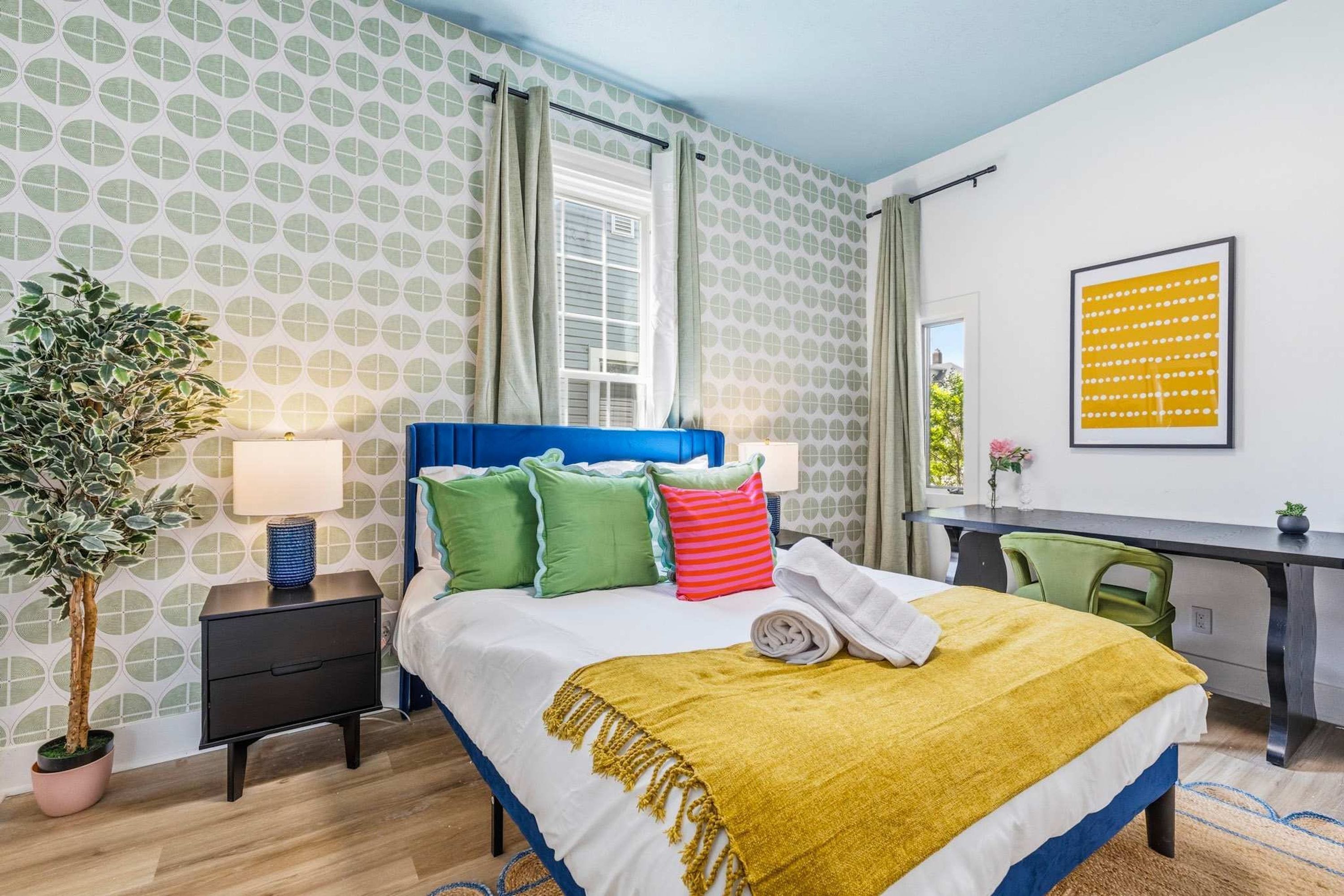 A hotel room with a bed, bedside table, lamp, potted plant, desk, chair, and a framed picture on the wall.