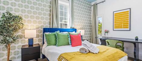 A hotel room with a bed, bedside table, lamp, potted plant, desk, chair, and a framed picture on the wall.