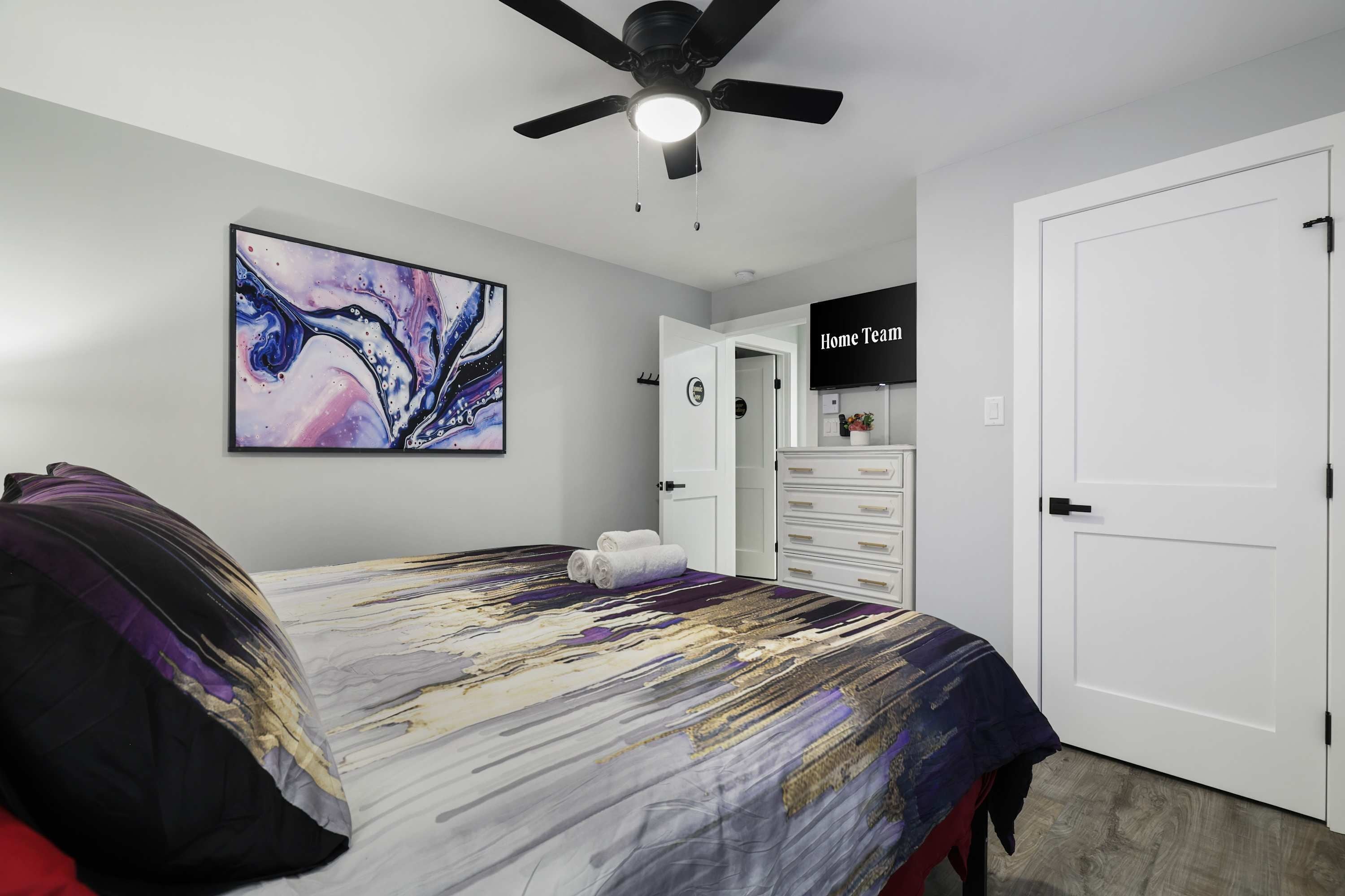 A bedroom with a bed, a ceiling fan, a wall-mounted TV, and a door.