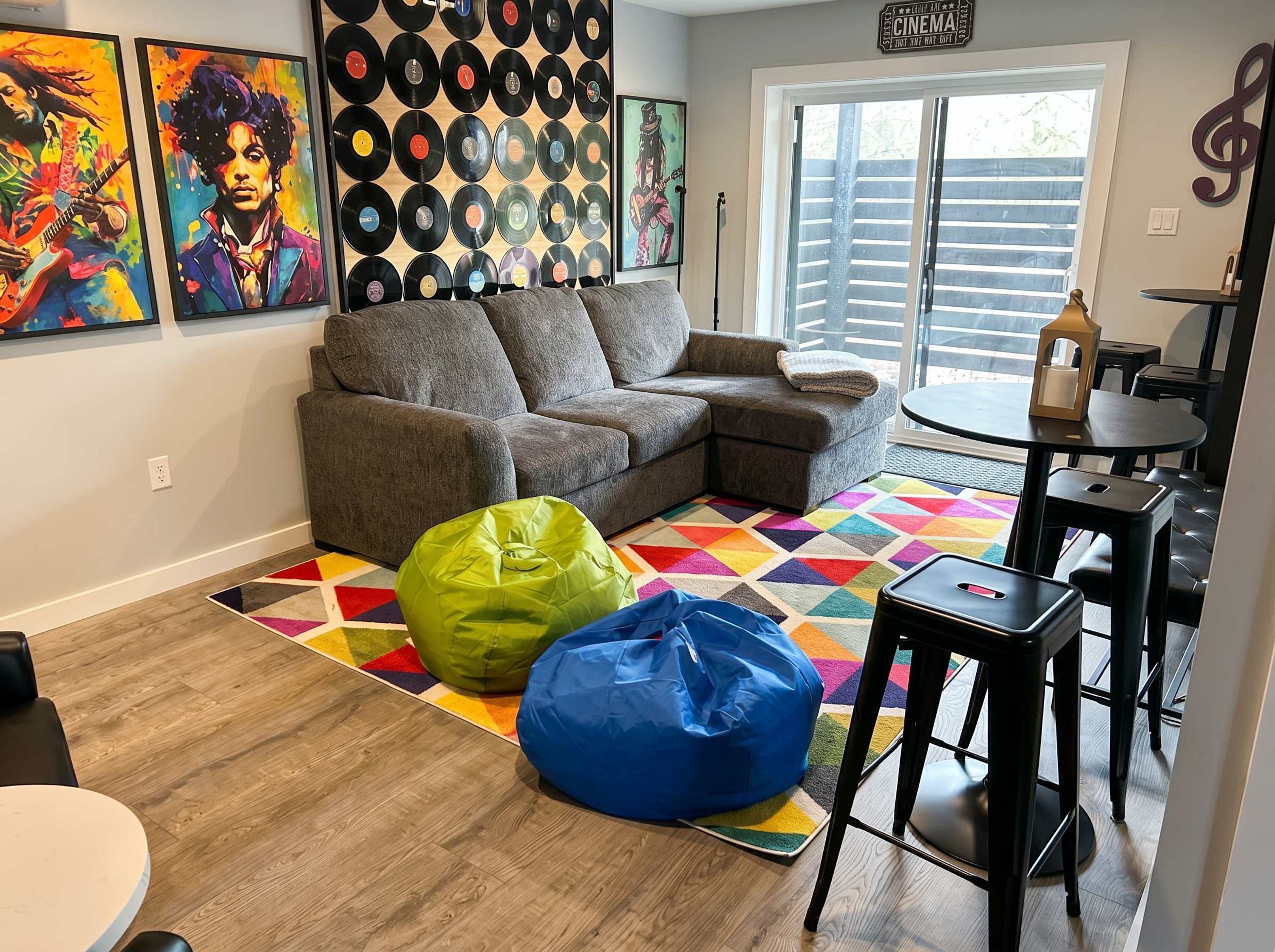 A living room with a gray sectional sofa, colorful geometric rug, green and blue bean bags, a wall of vinyl records, and a large window with blinds.