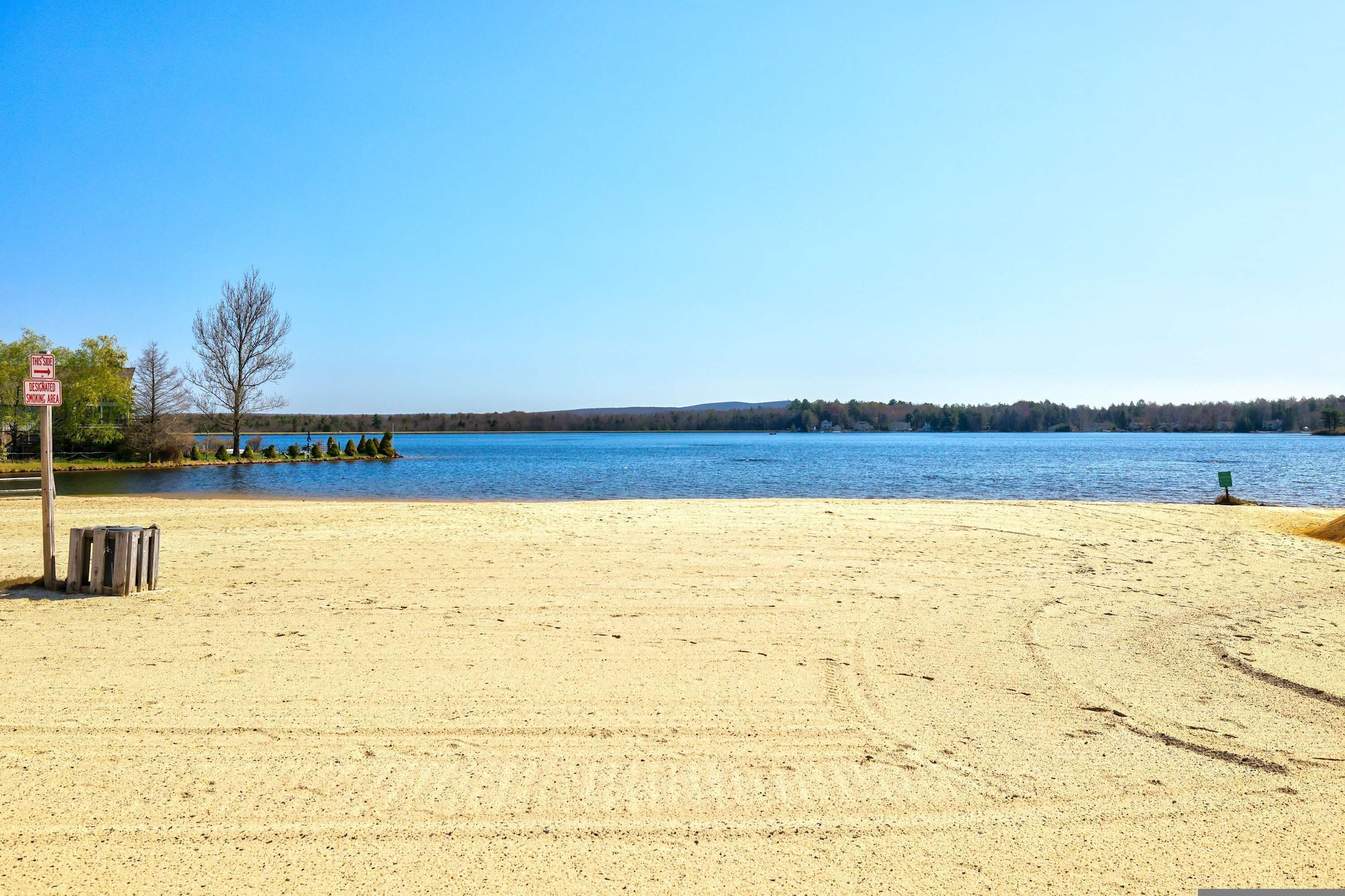 A sandy beach with a clear blue lake, a designated smoking area sign, and a row of trees.