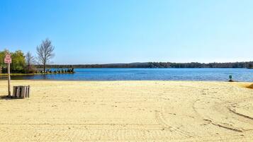 A sandy beach with a clear blue lake, a designated smoking area sign, and a row of trees.