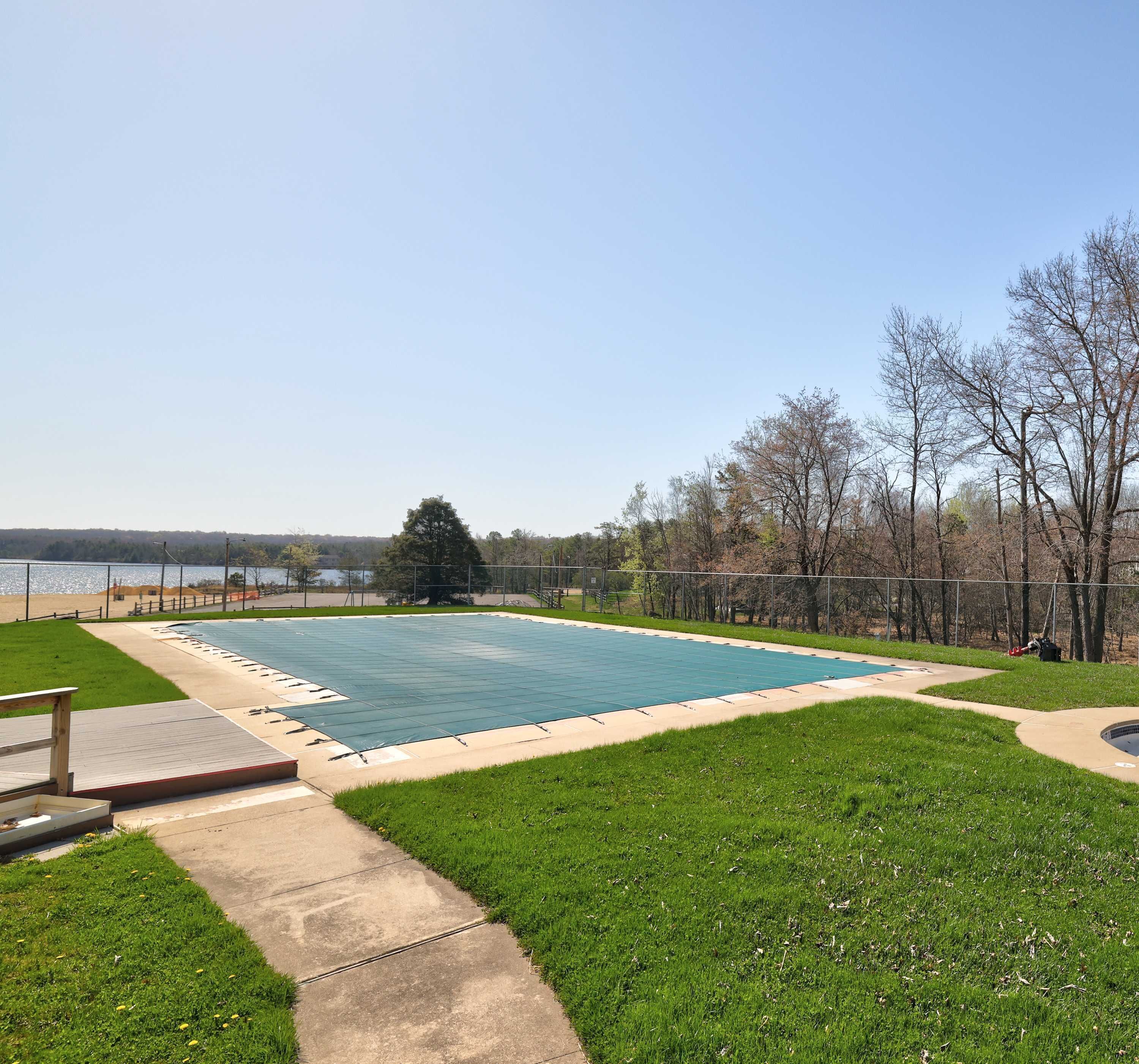 A rectangular swimming pool with a surrounding deck, a grassy area, and a view of a body of water and trees in the background.