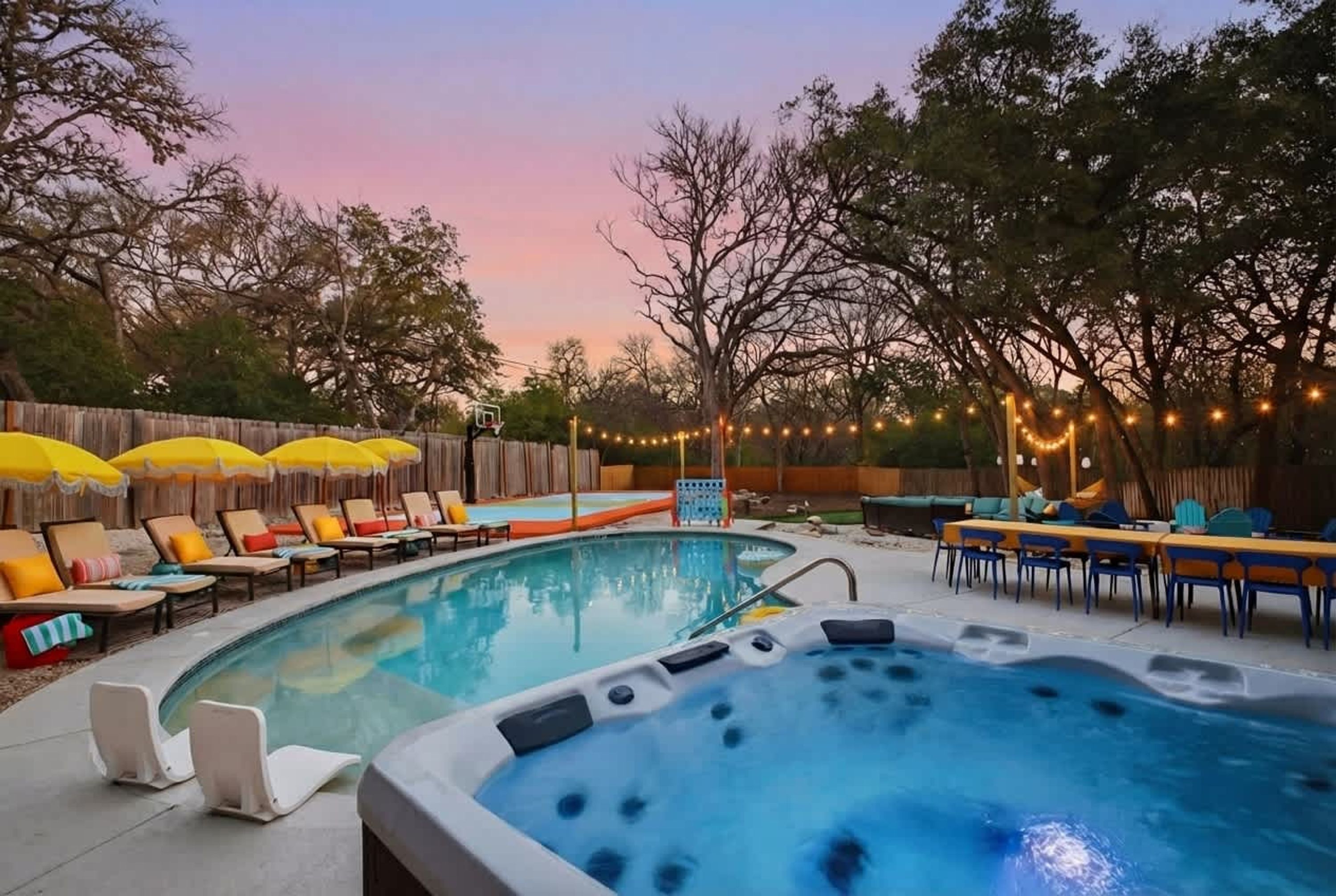 A backyard with a swimming pool, a hot tub, lounge chairs, and string lights.