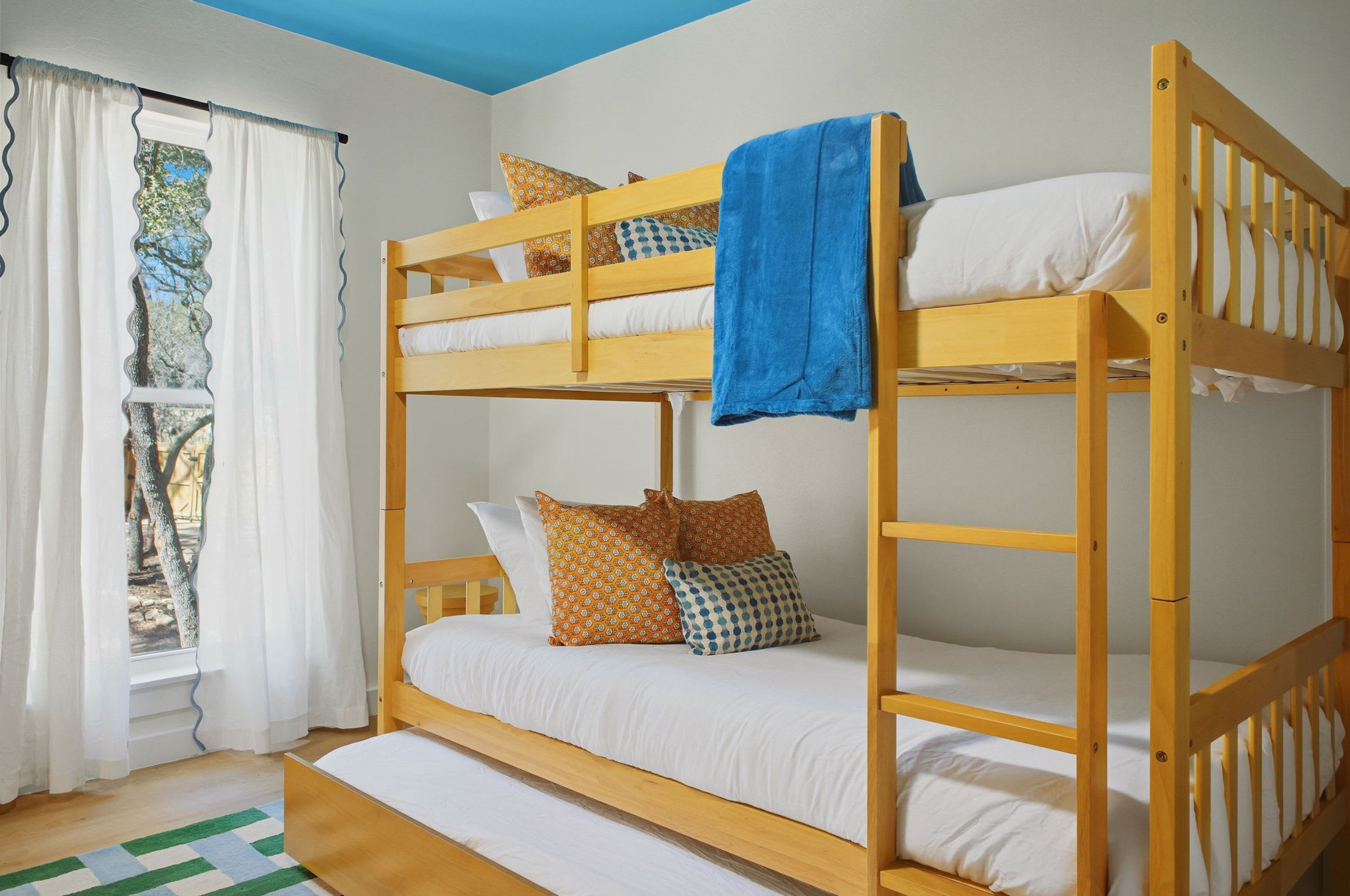 A bunk bed with white bedding and patterned pillows, a blue towel, and a window with white curtains.