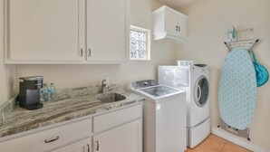 A compact kitchen with a sink, a coffee maker, a washing machine, and a window with blinds. - Sunrise Beach Villa Beach Views Private Elevator (Port Aransas)