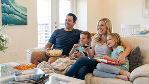 A family sitting on a couch with a bowl of popcorn and a remote control, in a room with a painting on the wall. - Sunrise Beach Villa Beach Views Private Elevator (Port Aransas)