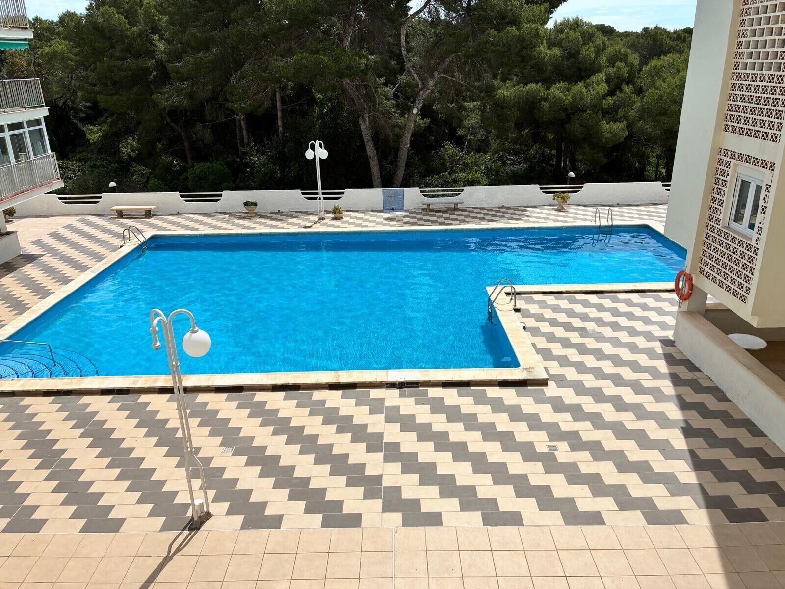 A swimming pool with a chevron-patterned deck, surrounded by trees and a building with a balcony.