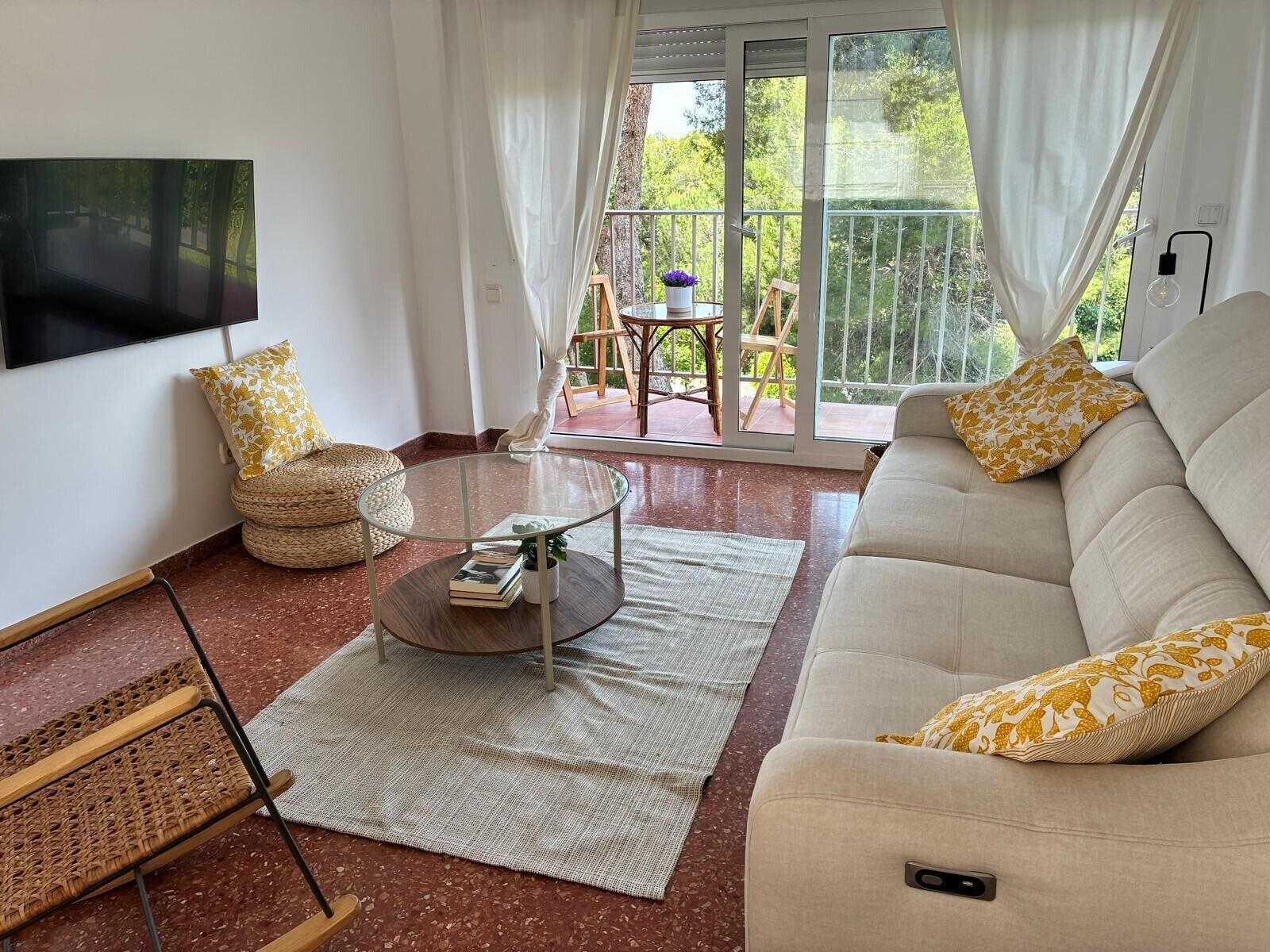 A living room with a sofa, a glass coffee table, a wicker chair, a flat-screen TV mounted on the wall, and a sliding glass door leading to an outdoor area with a table and chairs.
