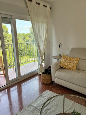 Ein Zimmer mit einer Couch, einem kleinen Tisch mit Lampe, einem Korb und Blick auf einen Balkon mit Geländer.