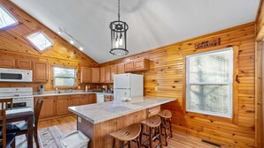 A cabin-style kitchen with wooden walls, a central island with stools, a microwave, a refrigerator, and a hanging light fixture. - Cozy Cabin in Mountains Firepit Views Games (Sautee Nacoochee)