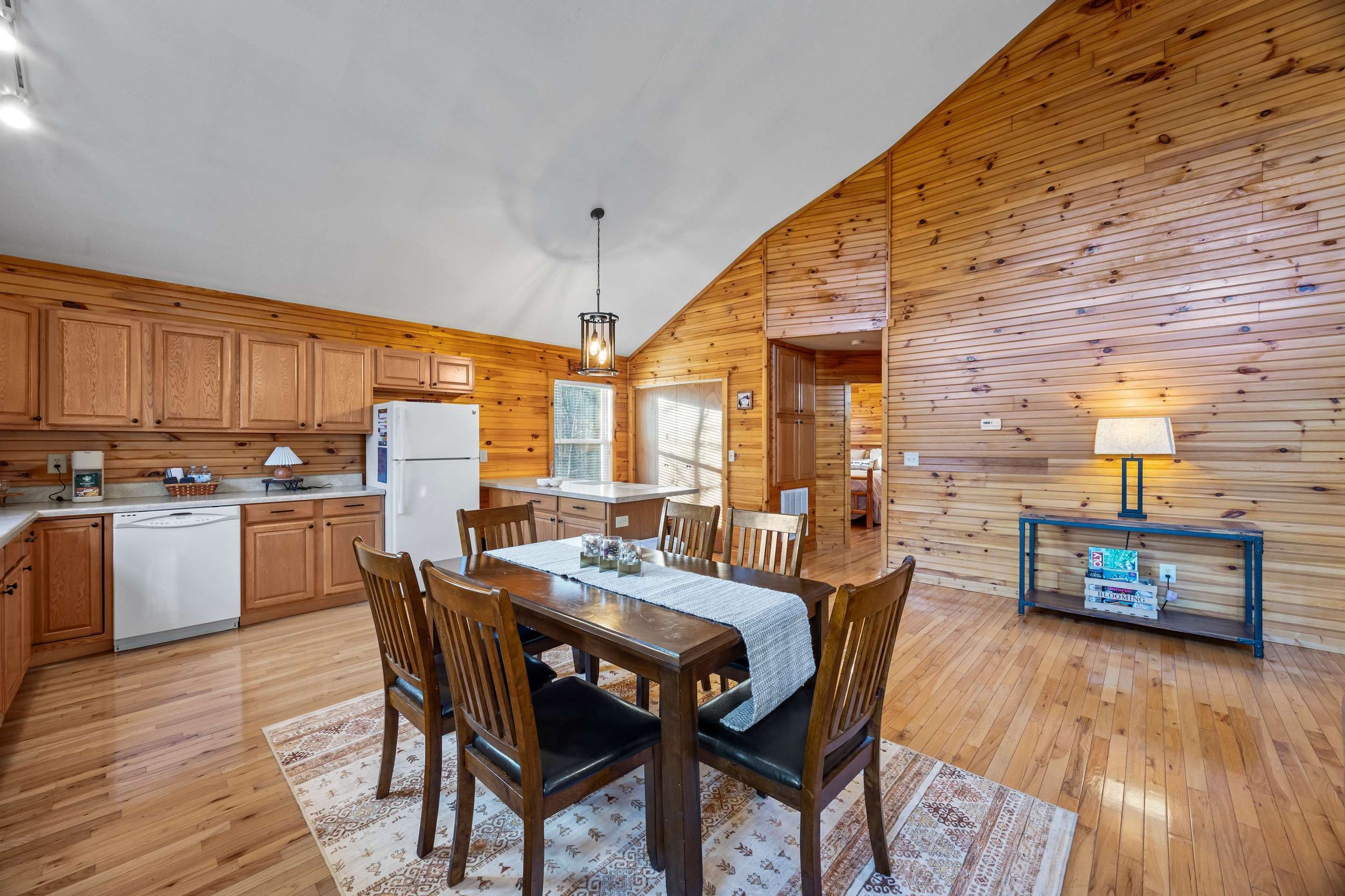 A wooden interior with a dining table, chairs, a kitchen area, and a small side table with a lamp.