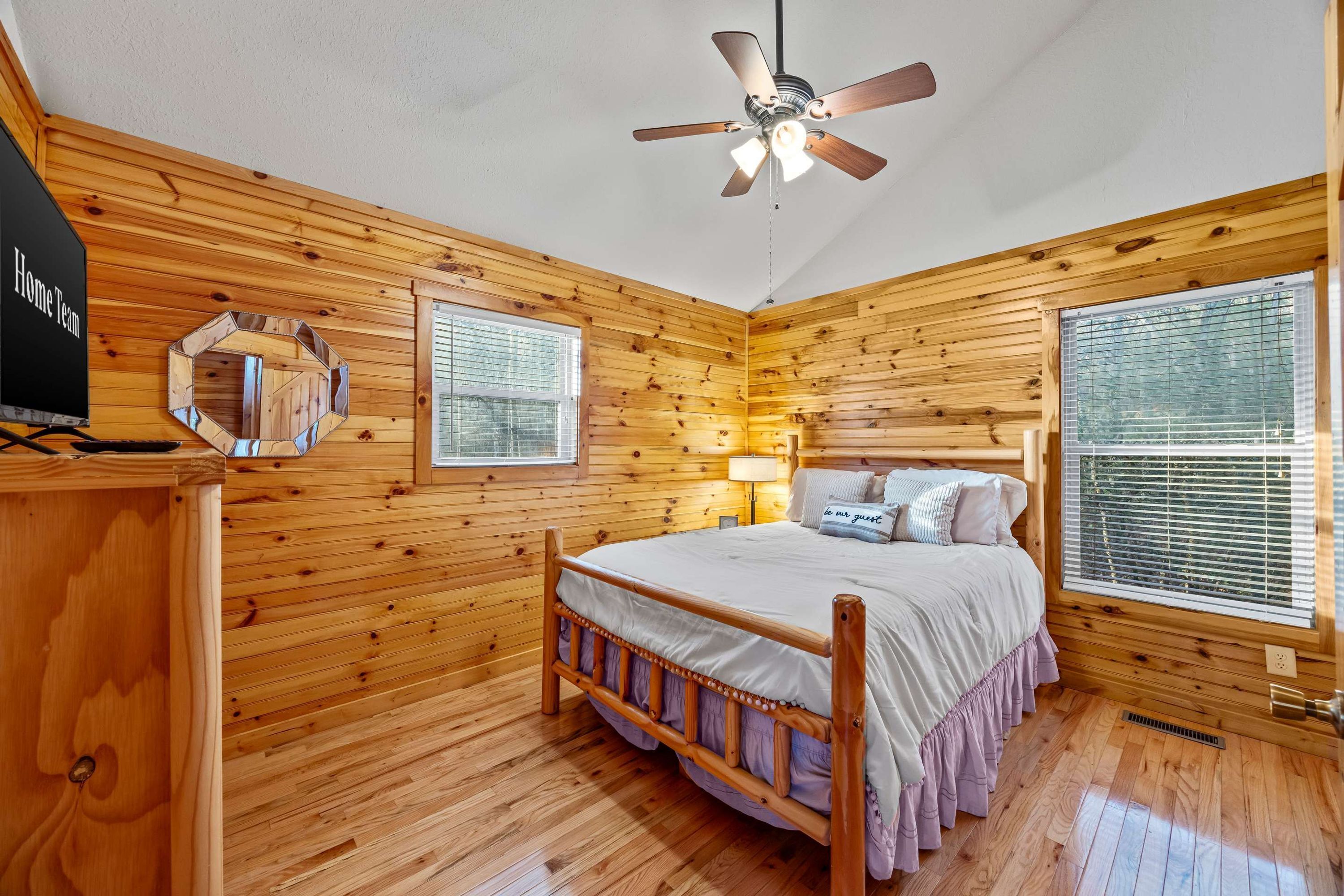 A wooden cabin bedroom with a bed, a ceiling fan, a nightstand, a mirror, and a window with blinds.