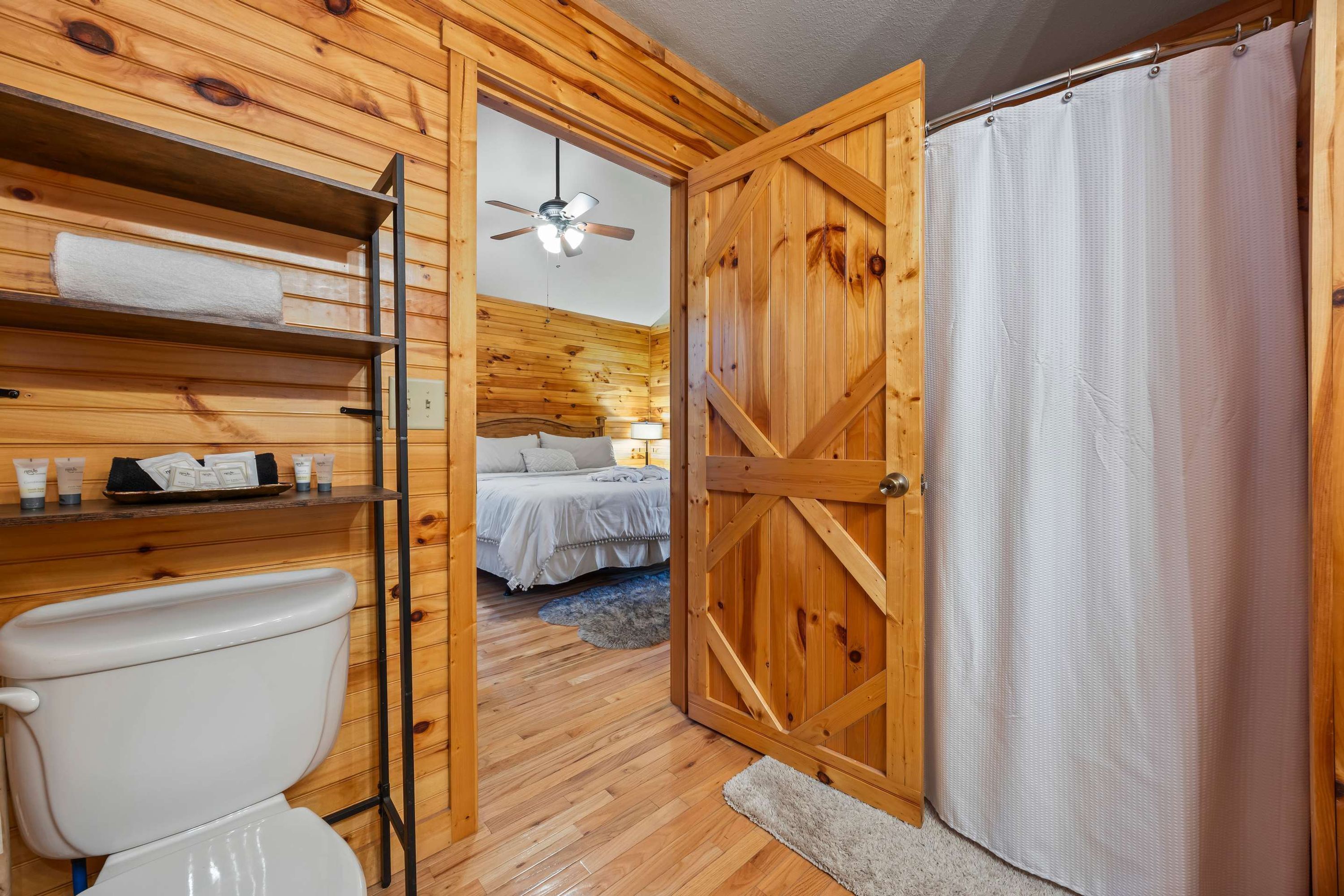 A wooden interior with a toilet, a shelf with toiletries, a bed with a nightstand, and a shower area.