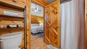 A wooden interior with a toilet, a shelf with toiletries, a bed with a nightstand, and a shower area.