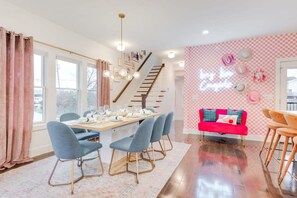 Comedor con una mesa de madera dispuesta para cuatro, sillas azules, una escalera y una pared rosa con un letrero de neón.