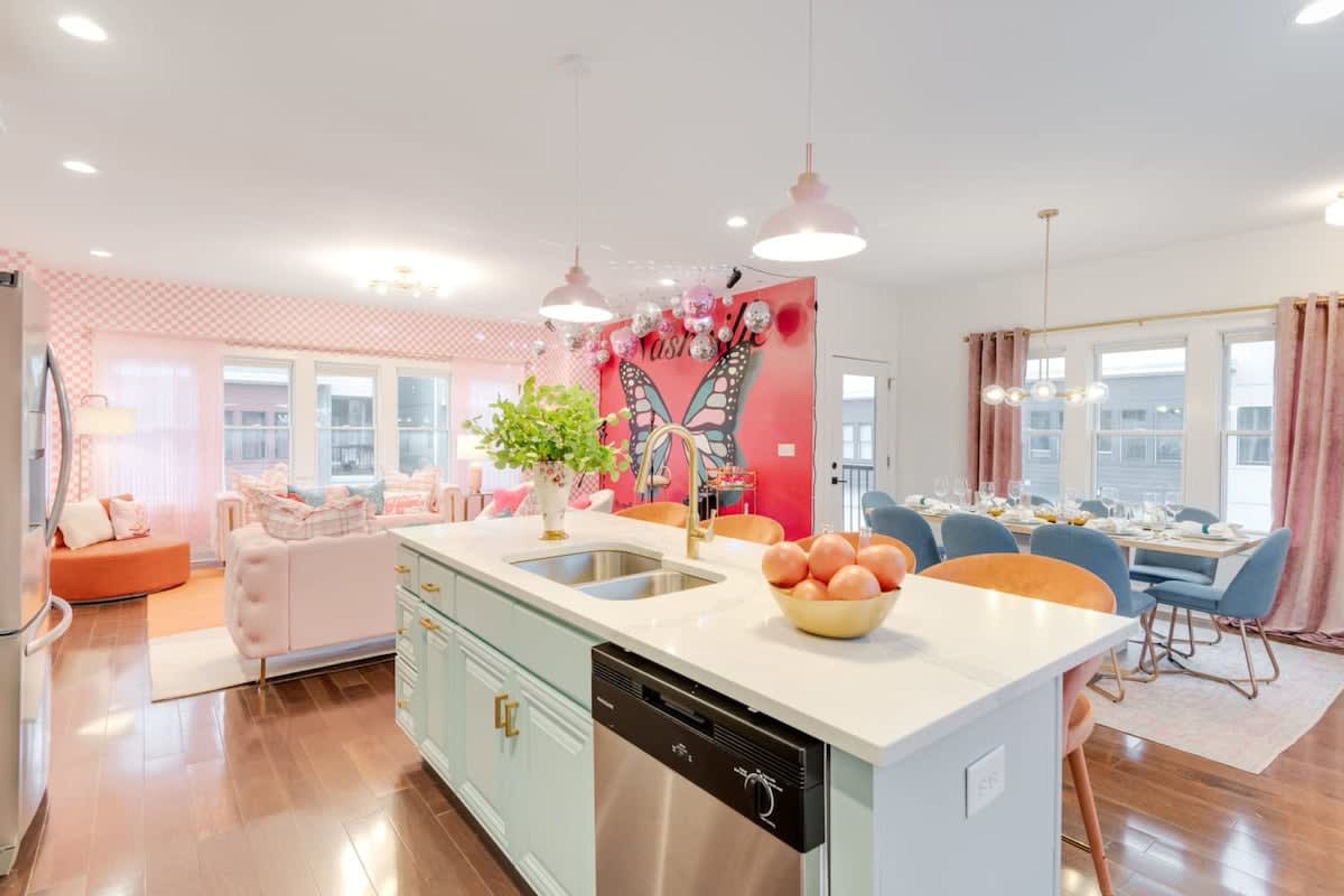 A modern kitchen with a central island, a dining area with blue chairs, and a living space with a pink sofa.