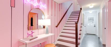 A pink-themed hallway with a neon sign, a staircase, and a mirror.