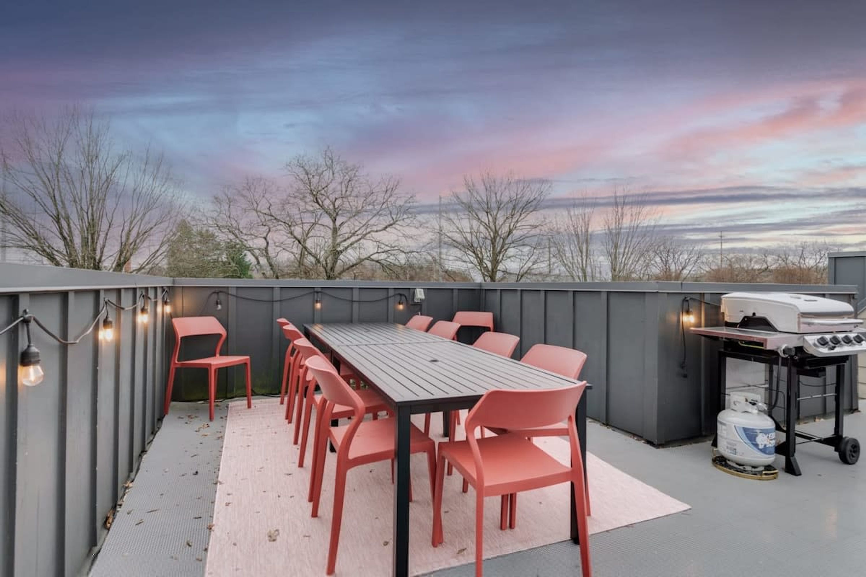 Une terrasse extérieure avec une table et des chaises, un barbecue et une vue sur les arbres et le coucher du soleil.