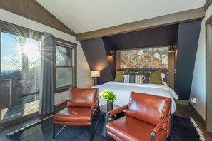 Quarto de hotel com uma cama grande, duas poltronas de couro, uma mesa pequena, um vaso de plantas, uma obra de arte de pedra na parede e uma janela com cortinas.