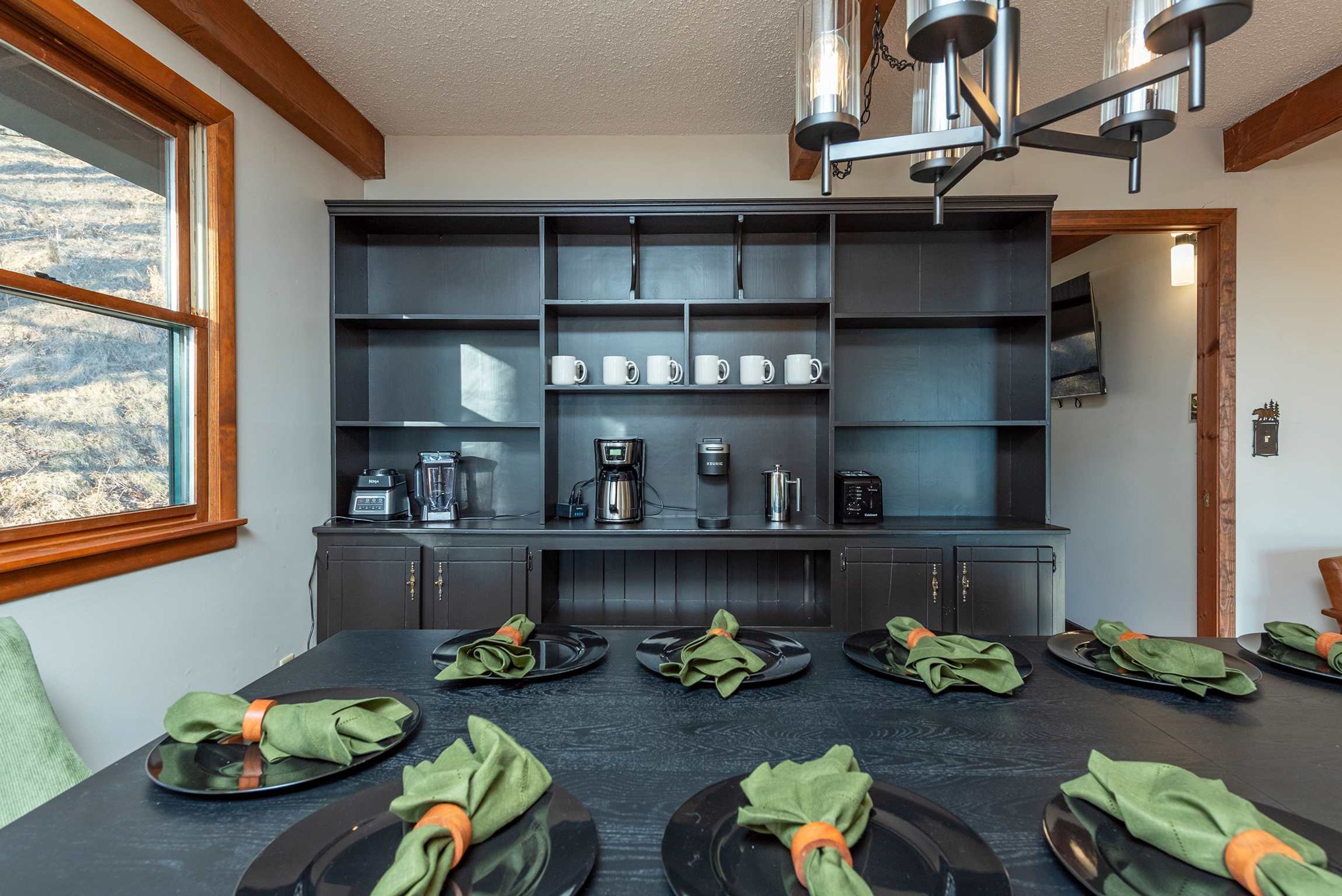 A modern kitchen with built-in appliances, a dining table set for six with green napkins, and a view of the outdoors through a window.