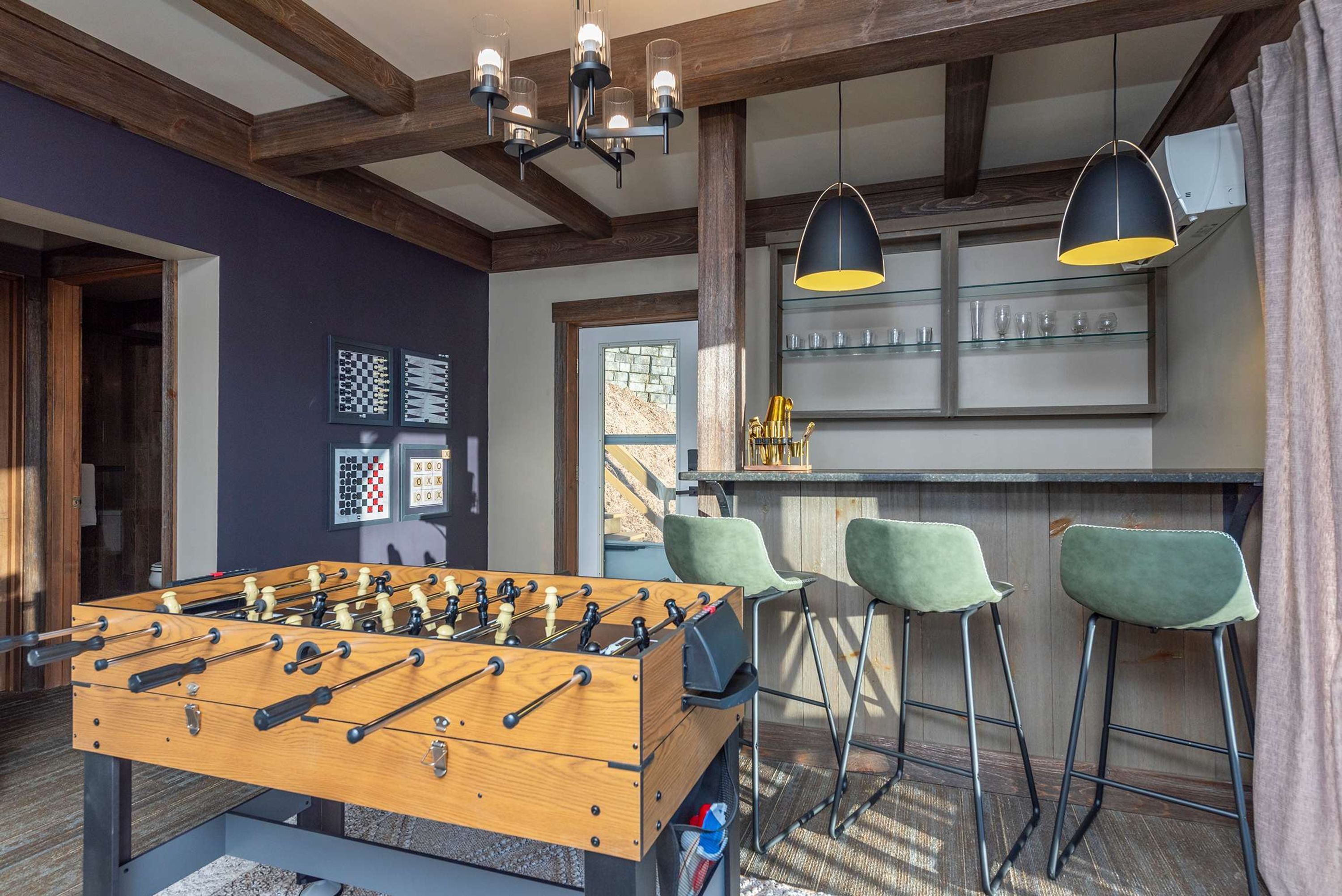 A room with a foosball table, bar area with stools, and a window with a view.