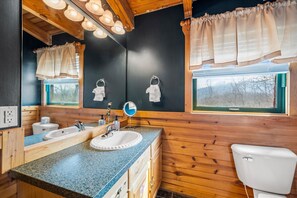 Ein Badezimmer mit Holzwänden, einer Granittheke, zwei Waschbecken, einer Toilette und einem Fenster mit Vorhängen.