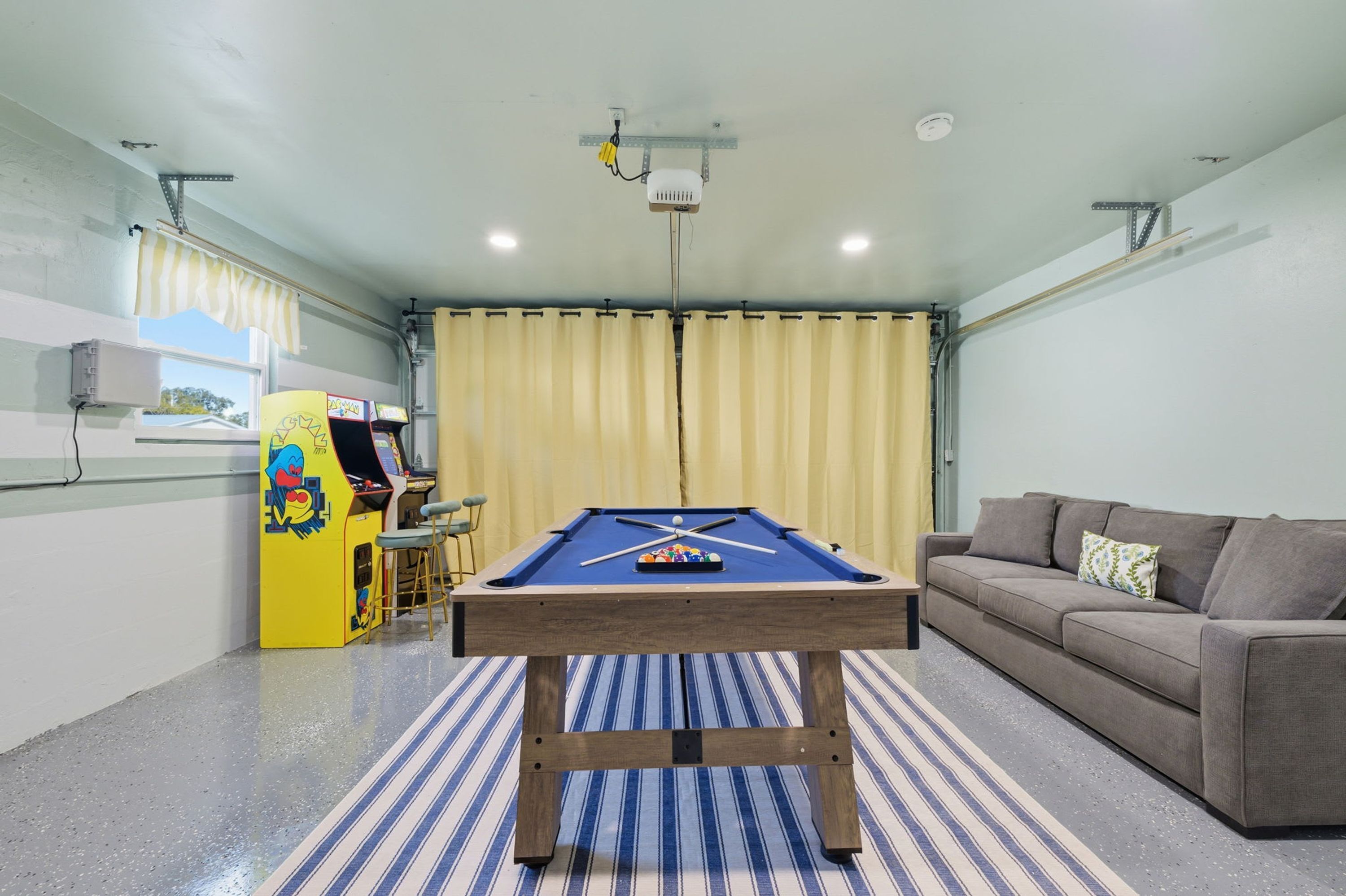 A pool table in a room with a sofa and a pool table, a ceiling fan, and a window with curtains.