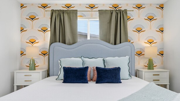 Quarto com cama, duas mesas de cabeceira, uma janela com cortinas e papel de parede estampado.