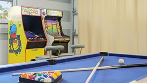 Uma mesa de bilhar com bolas e tacos, máquinas de jogos eletrônicos e uma janela com vista.