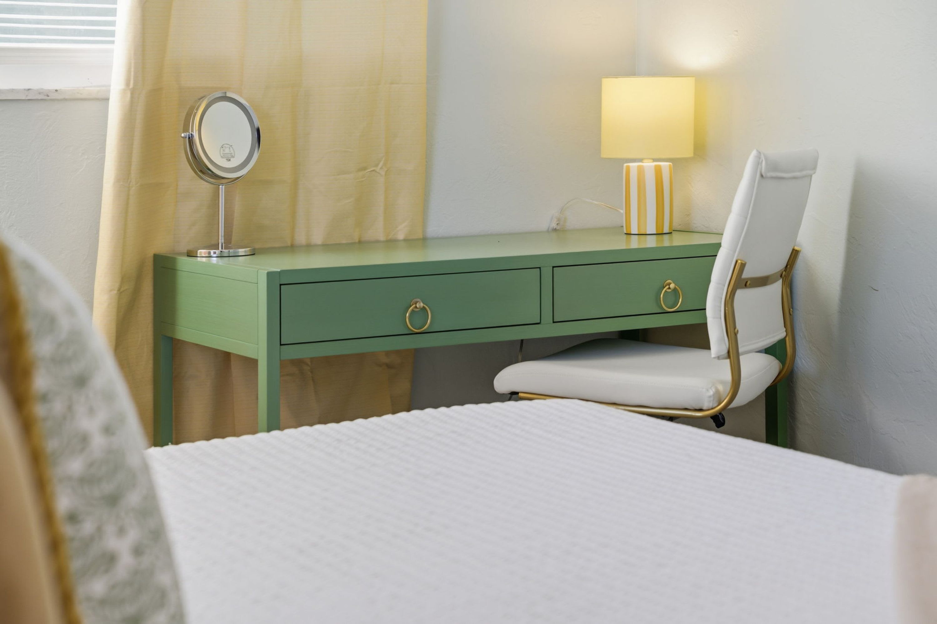 A room with a bed, a green desk with drawers, a chair, a bedside table with a lamp, and a window with curtains.