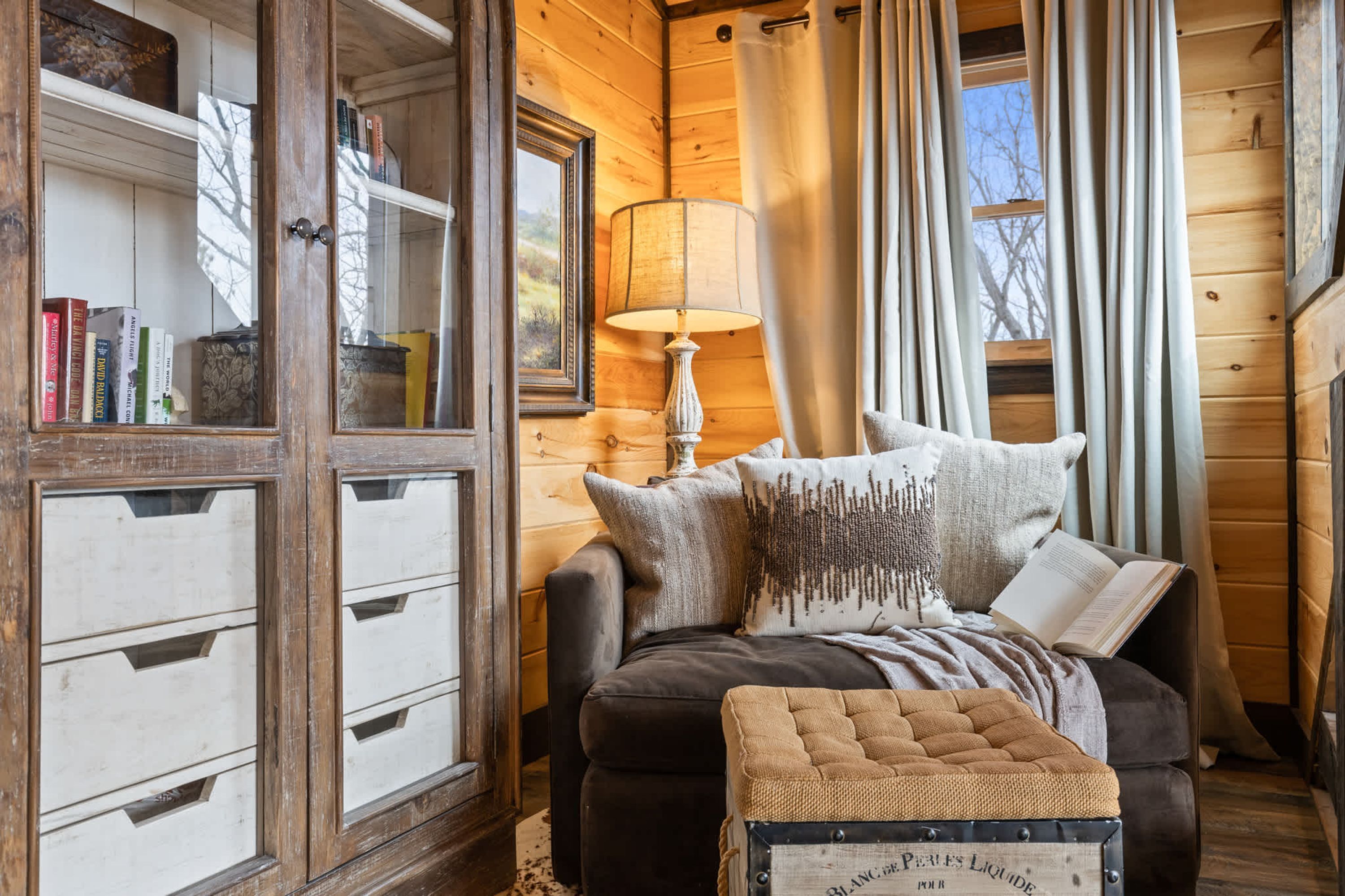 A cozy log cabin room with a wooden bookshelf, a comfortable sofa with pillows, a lamp, and a window with curtains.