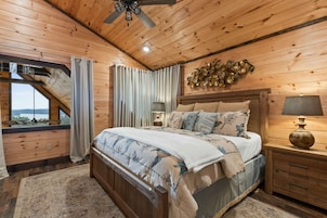 Un dormitorio de cabaña de madera con una cama, mesitas de noche, un ventilador de techo y vistas por una ventana.