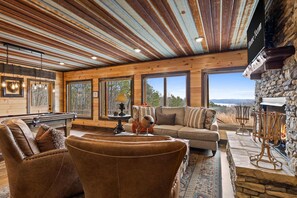 A spacious living room with a pool table, leather chairs, a stone fireplace, and large windows offering a view of the outdoors. - Epic Mountain Views Hot Tub Games and Firepit (Ellijay)