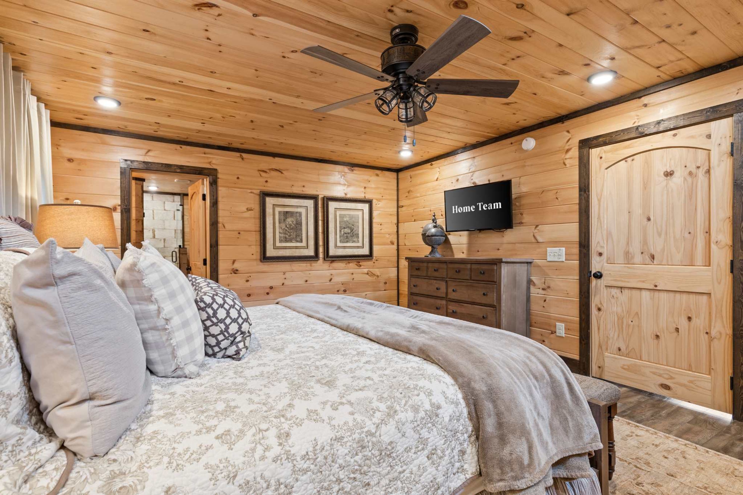 A bedroom with a bed, pillows, a ceiling fan, a dresser, a TV, and wooden walls.