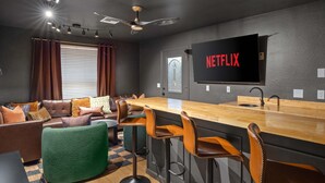 A modern living room with a bar area, leather sofa, and a large flat-screen TV displaying 'NETFLIX'. - Epic Family Getaway Speakeasy Pool Basketball (Glendale)